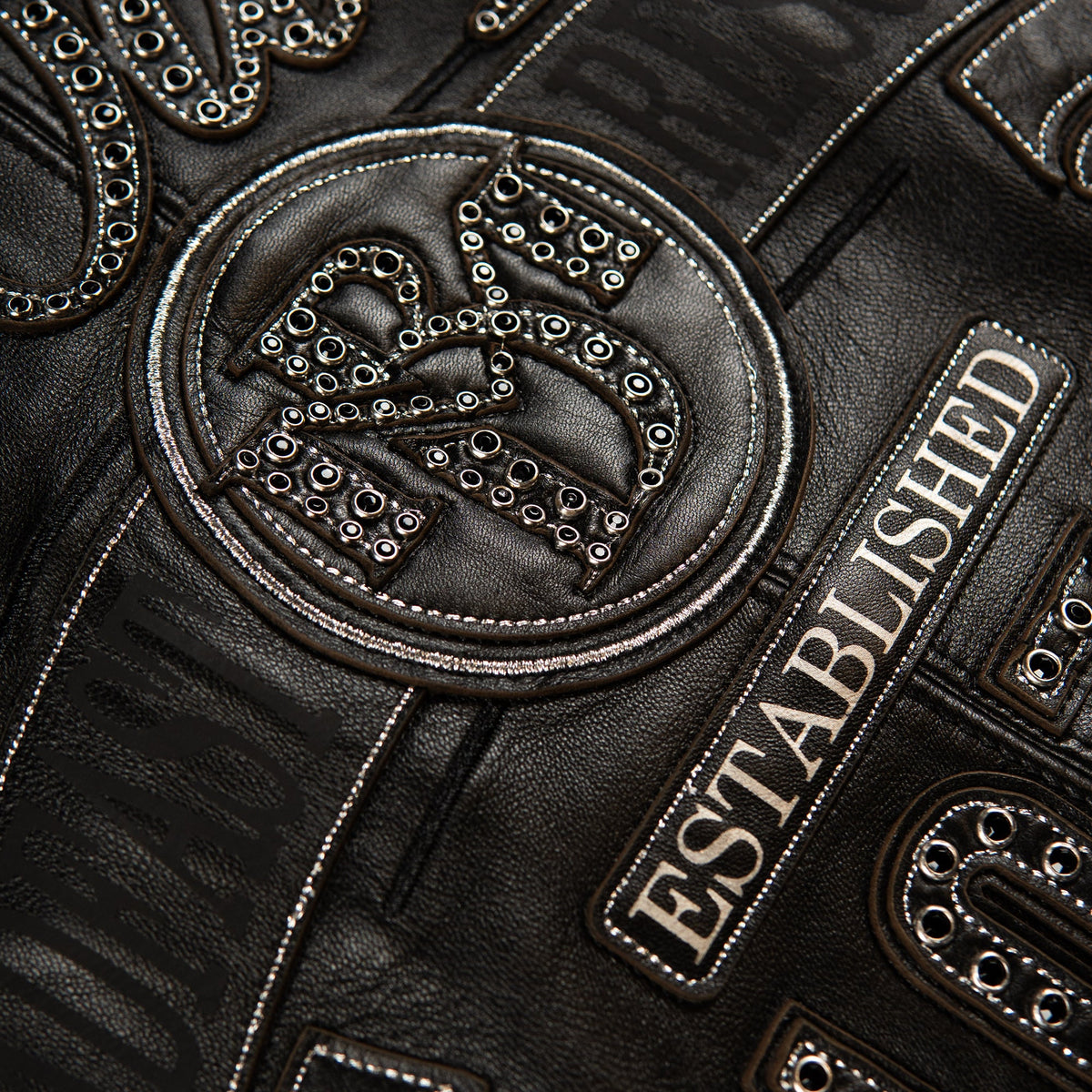 Close-up of a black leather jacket featuring an embroidered circular patch with a stylized 'M' and 'B' monogram, embellished with silver studs and white contrast stitching, alongside a rectangular patch reading 'ESTABLISHED' in beige lettering.