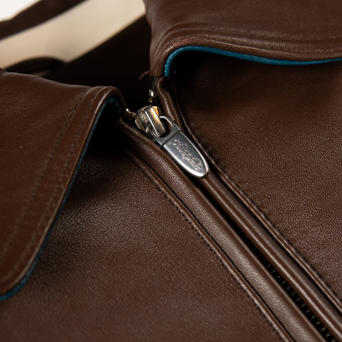 Close-up of a brown leather jacket with a silver metal zipper and a small visible pelle logo on the zipper pull.