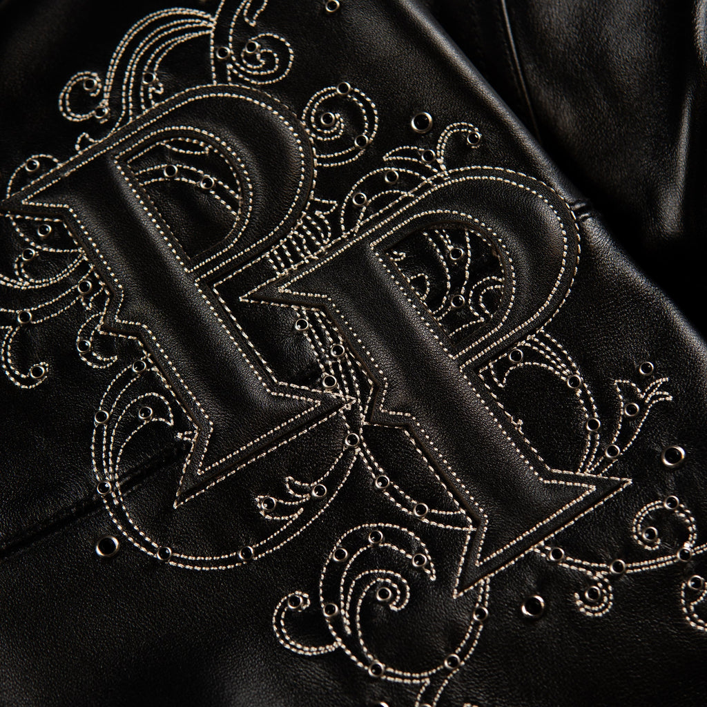 Close-up of a black leather jacket featuring an ornate, raised embroidered monogram logo with intricate white contrast stitching and decorative scrollwork.
