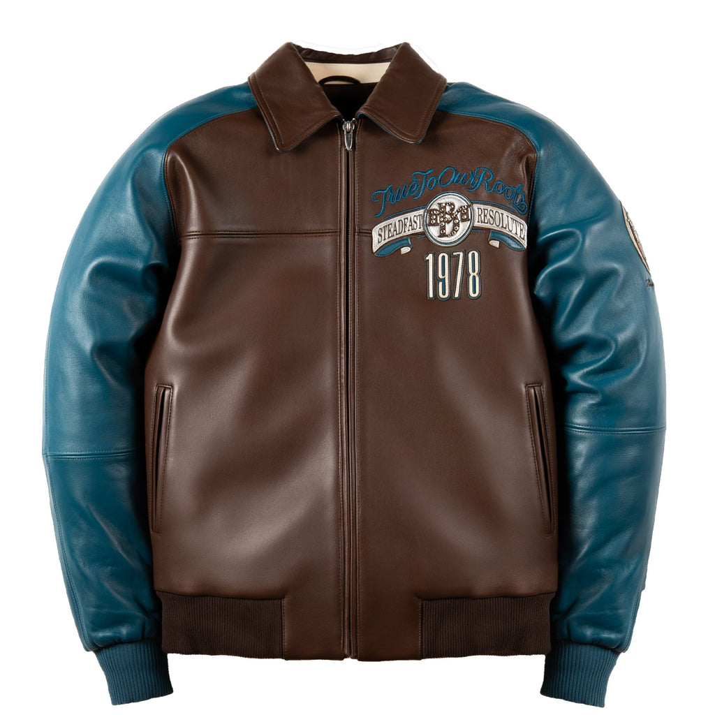 A brown and teal leather varsity-style jacket with a front zipper, ribbed collar, cuffs, and hem, featuring 'Trust No One 1978' embroidered on the left chest.