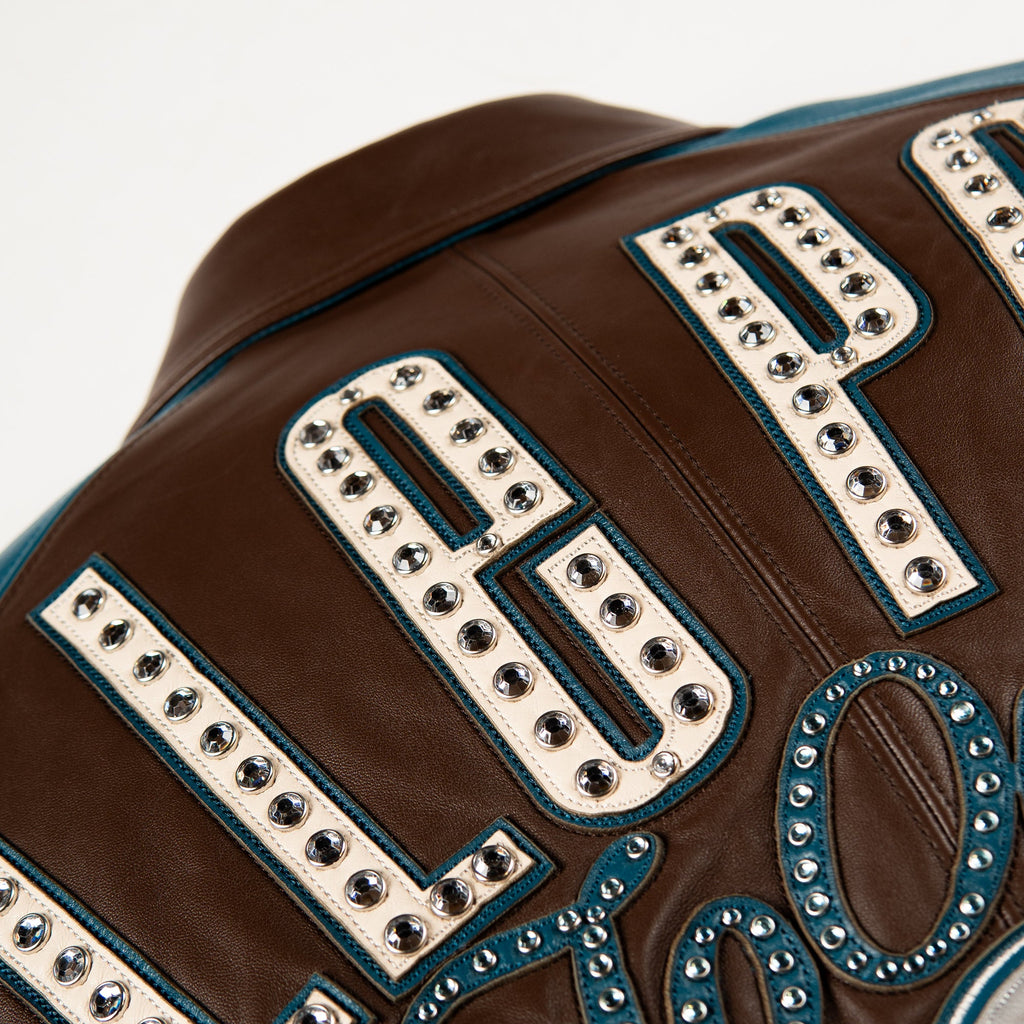 Close-up of a brown leather jacket featuring large cream-colored letter patches outlined with silver studs and teal stitching.