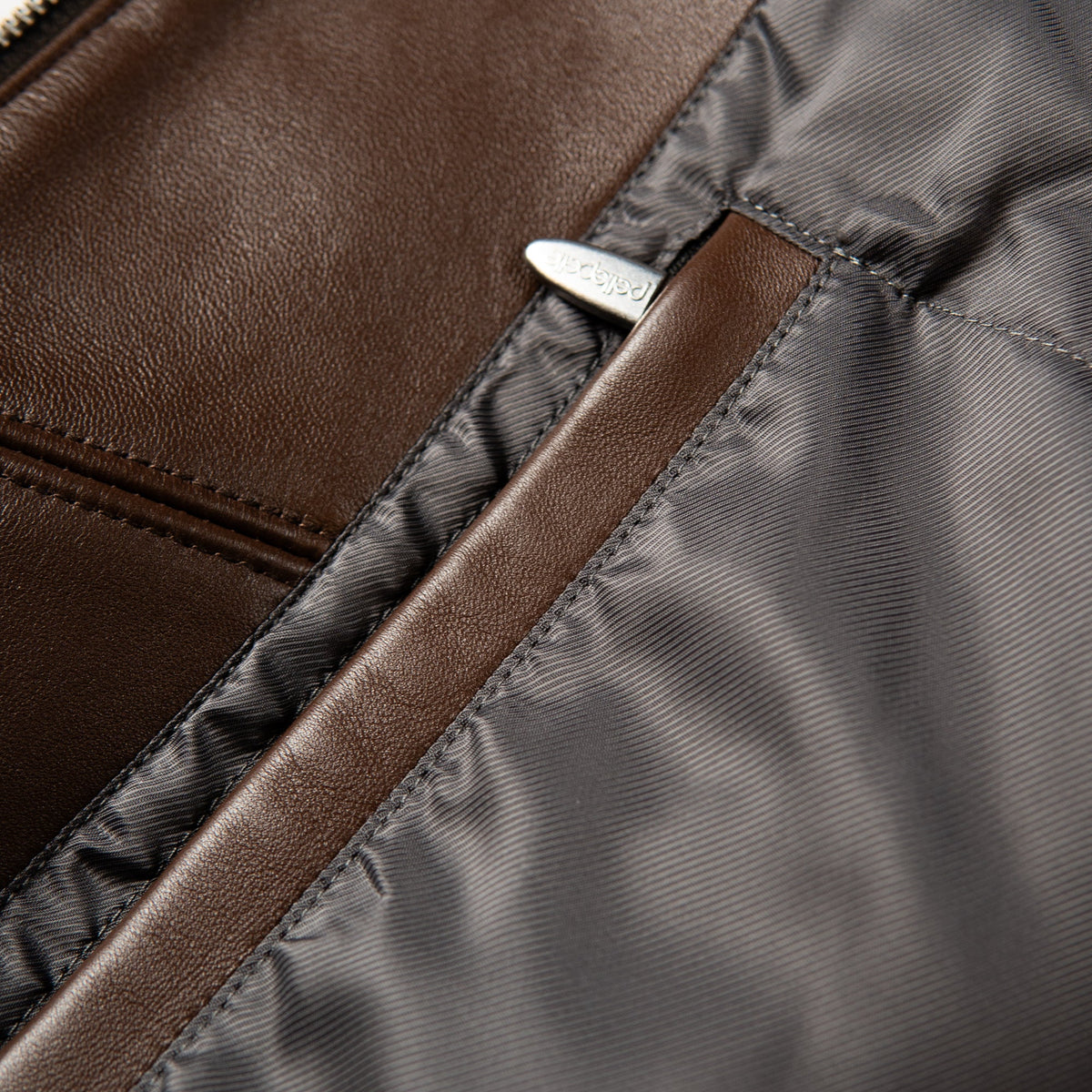 Close-up detail of a brown leather and dark gray nylon bag, showing a silver-tone metal zipper pull engraved with the brand name 'pelle'.