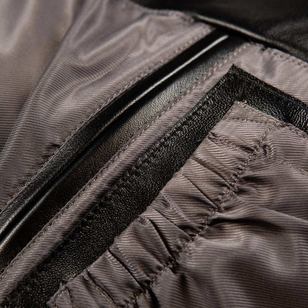 Close-up detail of a dark gray and black textured jacket, showing a zippered pocket with a black leather-look trim and a ribbed fabric lining.
