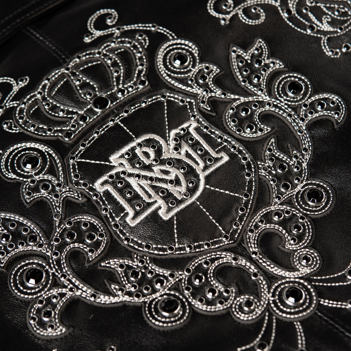 Close-up of a black leather jacket featuring intricate silver-toned embroidery and rhinestone embellishments, with a central shield motif and decorative scrollwork.