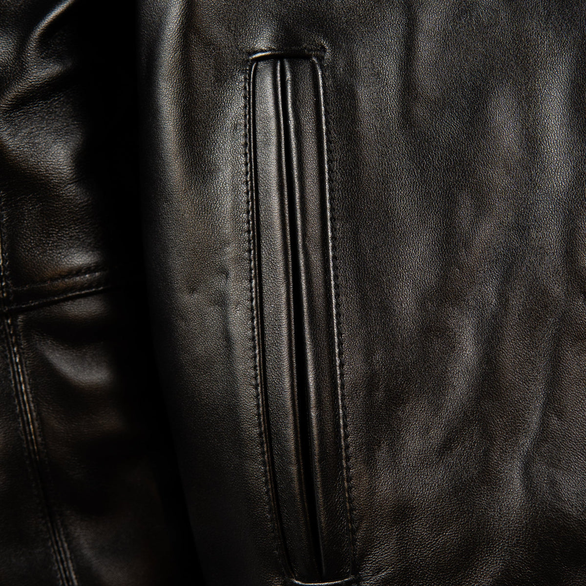 Close-up of a black leather jacket showing a vertical zippered pocket with precise stitching and a textured leather surface.