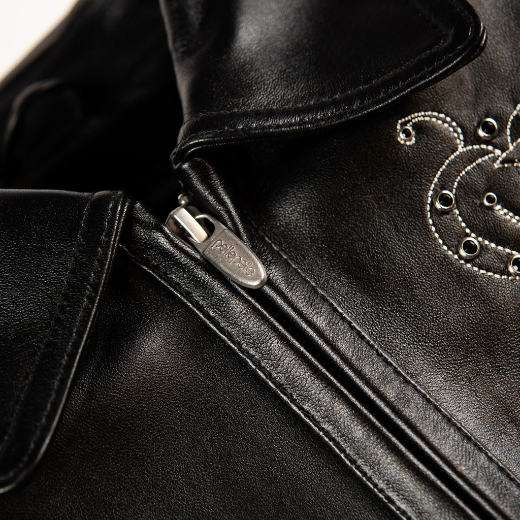 Close-up of a black leather jacket showing a silver zipper pull, fine stitching, and decorative silver grommet embroidery on the collar.