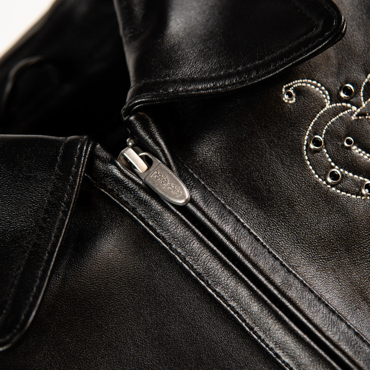 Close-up of a black leather jacket showing a silver zipper pull, fine stitching, and decorative silver grommet embroidery on the collar.