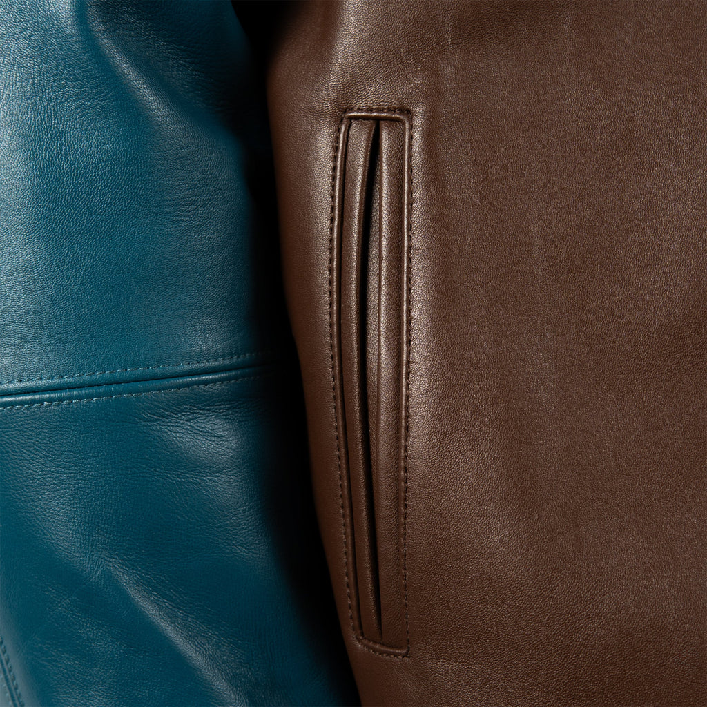Close-up of a brown leather jacket showing a vertical welt pocket and adjacent stitching, next to a teal blue leather section.