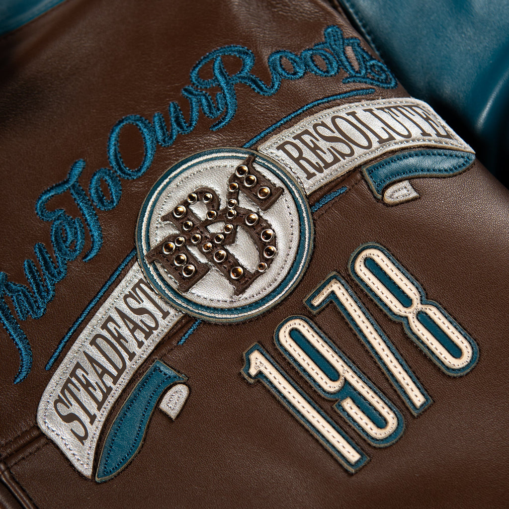 Close-up of a brown leather jacket featuring embroidered patches and text, including 'True To Our Roots', 'Steadfast Resolute', '1978', and a circular emblem with metallic studs.