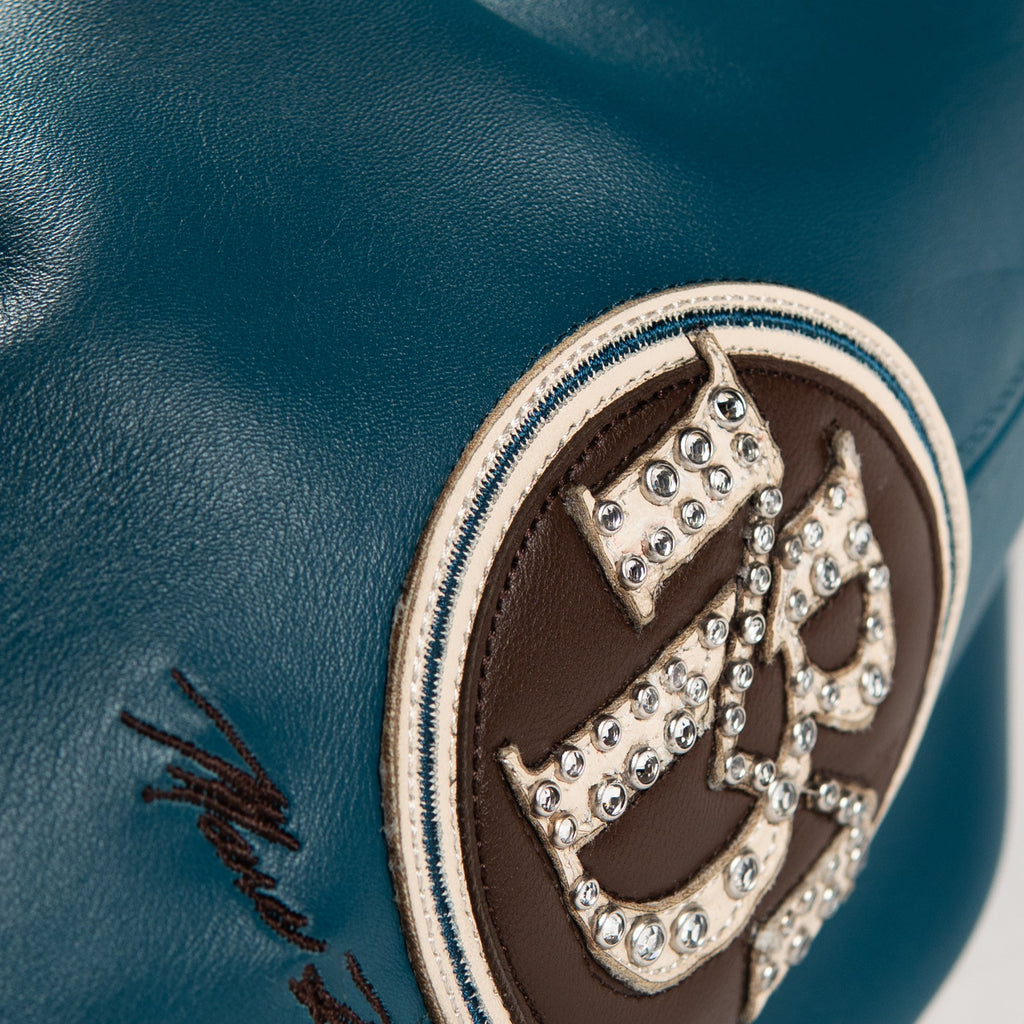 Close-up of a dark teal leather accessory featuring a circular brown patch with a stylized letter 'B' embellished with clear rhinestones, surrounded by a cream stitched border.