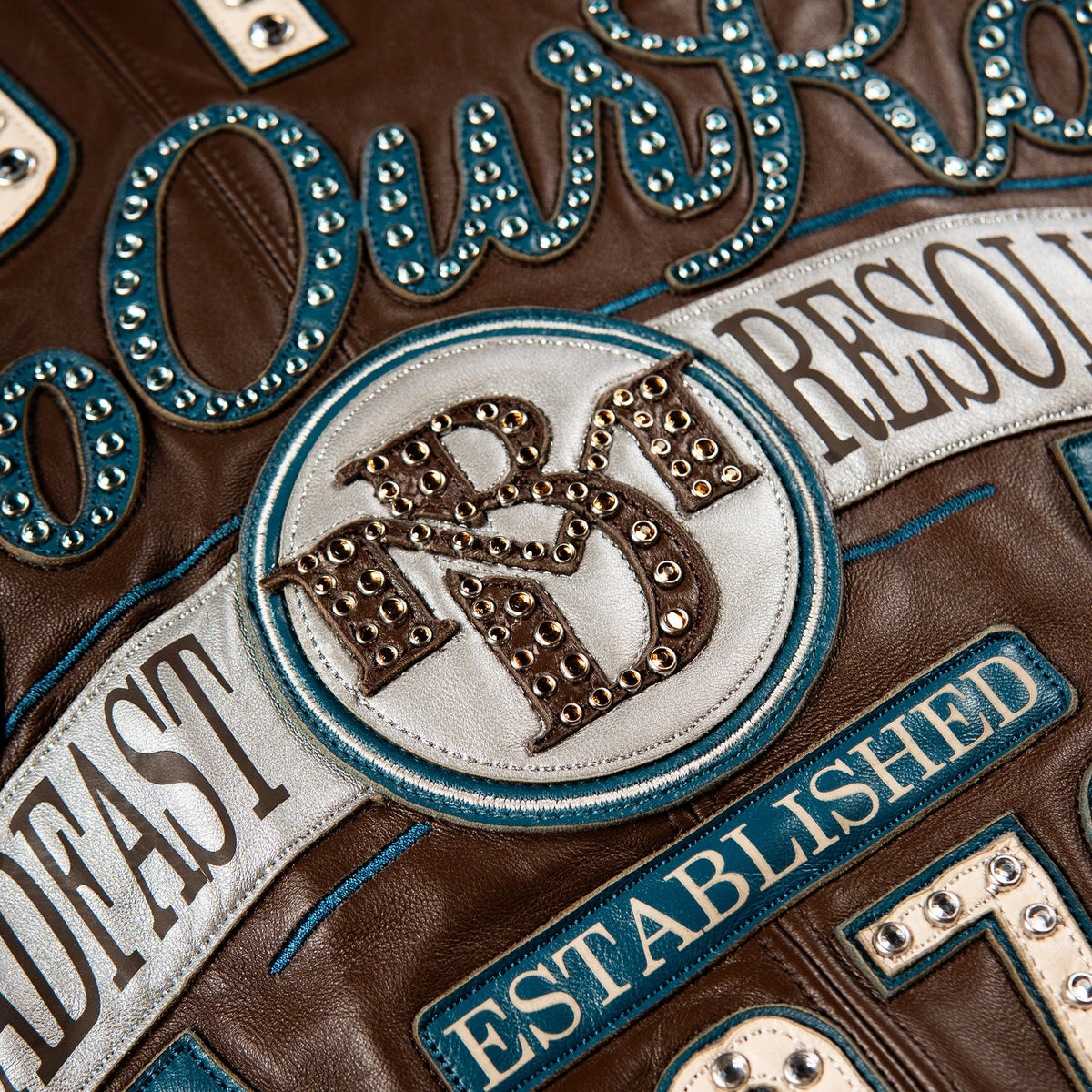 Close-up detail of a brown leather jacket featuring a circular white patch with brown embroidered letters outlined in rhinestones, surrounded by blue and white embroidered text and studs.