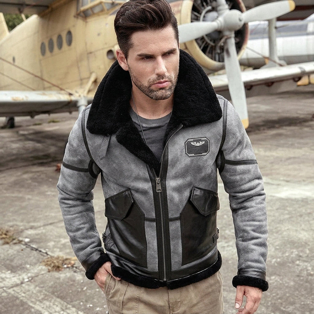 A man wearing a gray and black shearling-lined aviator jacket stands in front of an old airplane on a tarmac. The jacket features a plush black shearling collar, black trim, and a gray body with a black zipper and front pockets.