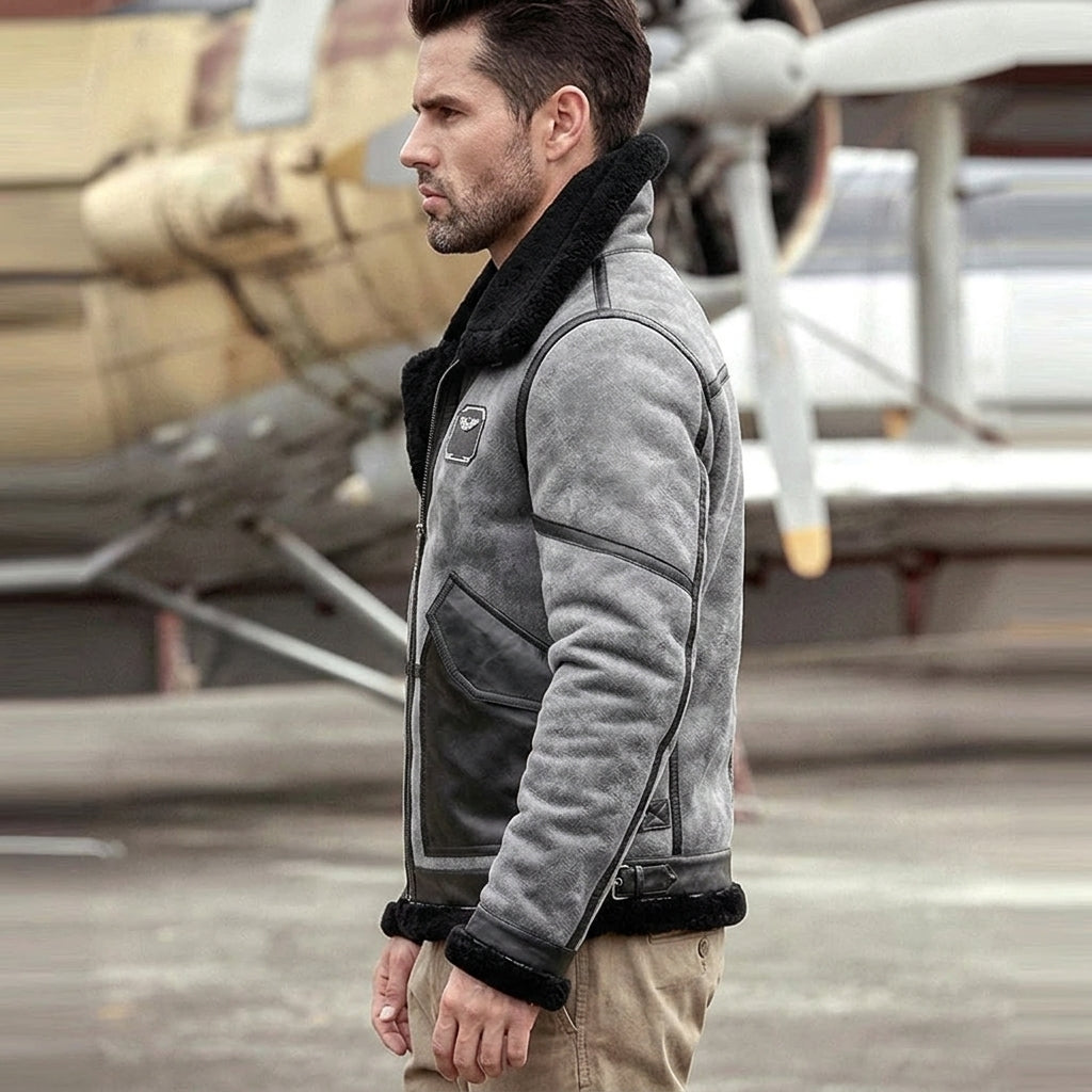 A man wearing a gray suede aviator jacket with black shearling lining stands in profile in front of a vintage airplane on an airfield.