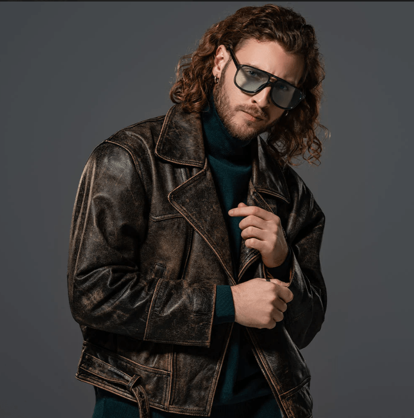 A man with long curly hair, a beard, and sunglasses wearing a distressed dark brown leather jacket over a dark teal turtleneck, posed against a solid dark gray studio background.