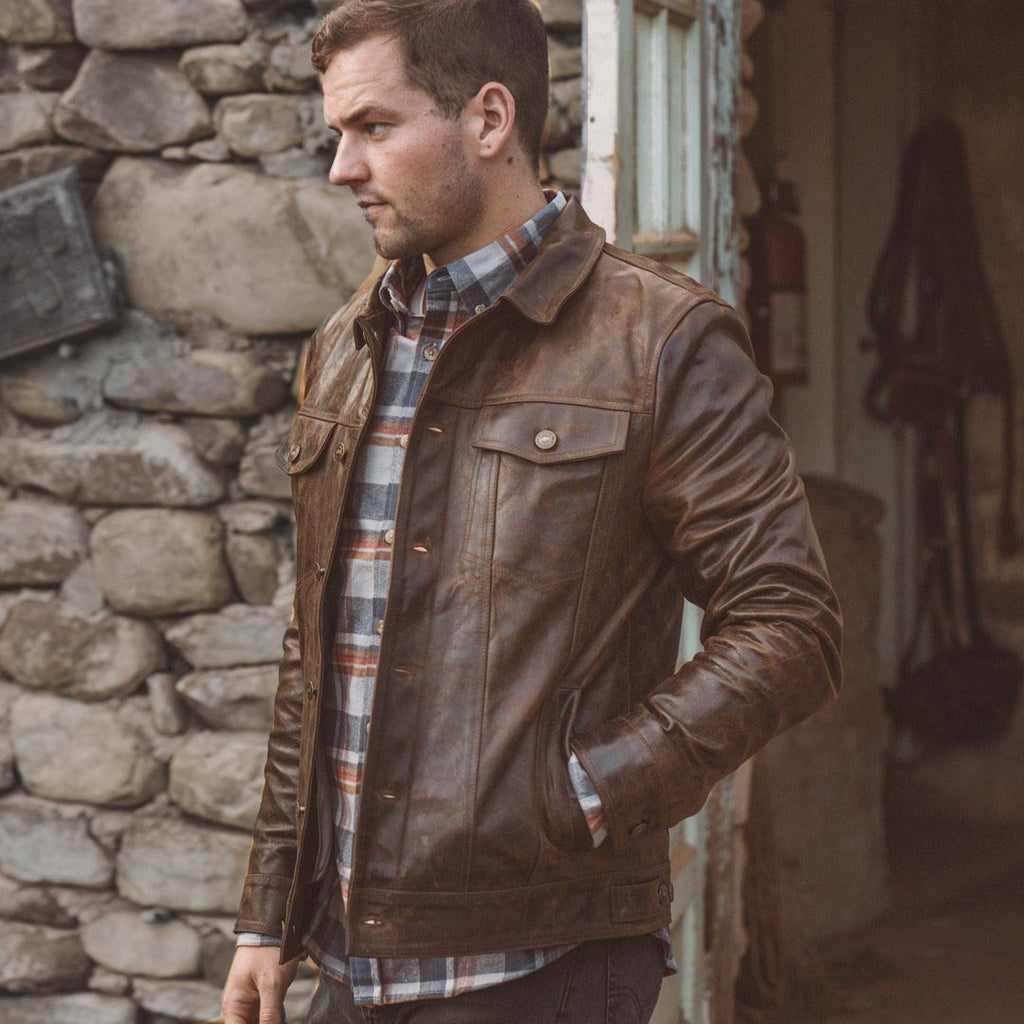 Man wearing a brown leather jacket and plaid shirt standing against a stone wall.