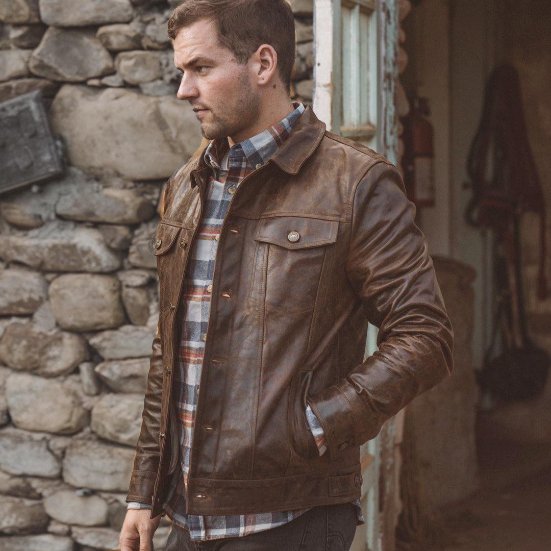 Man wearing a brown leather jacket and plaid shirt standing against a stone wall.