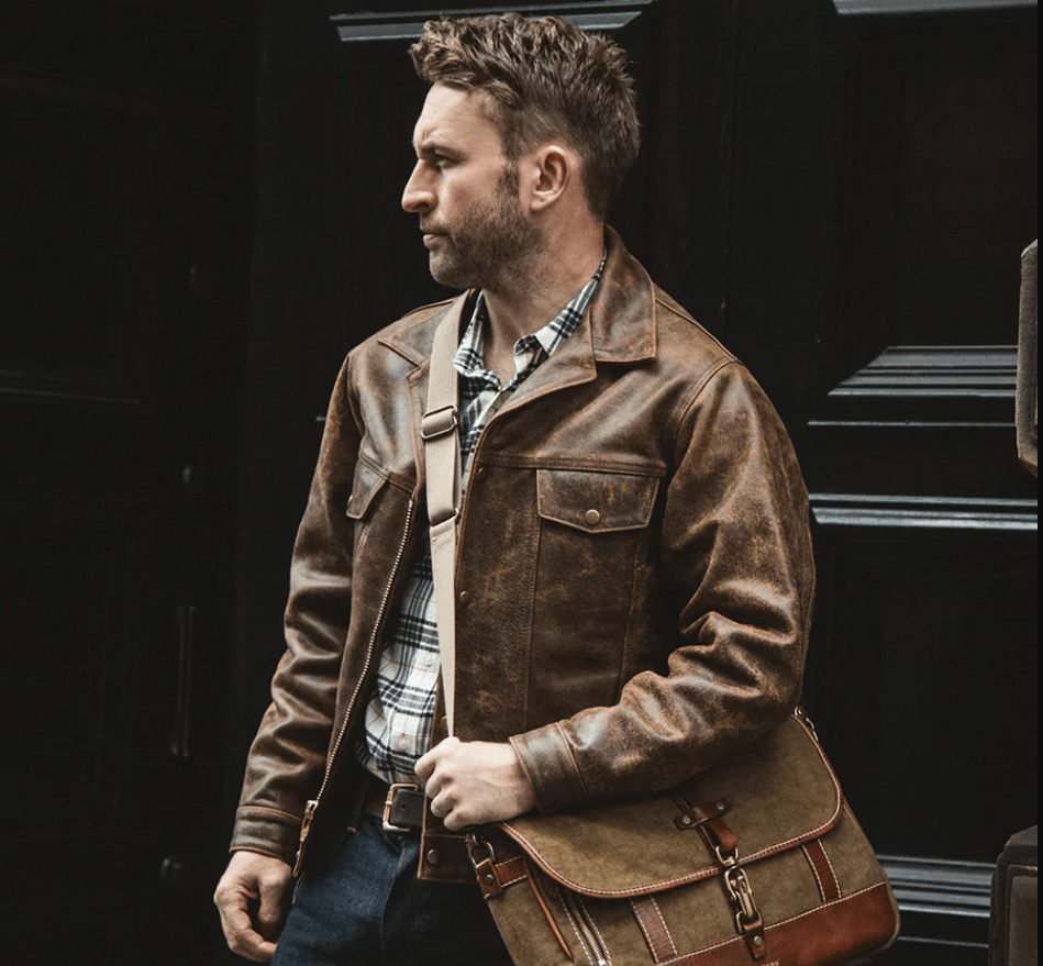 A man wearing a distressed brown leather jacket and carrying a matching brown leather messenger bag over his shoulder, standing against a dark wooden door.