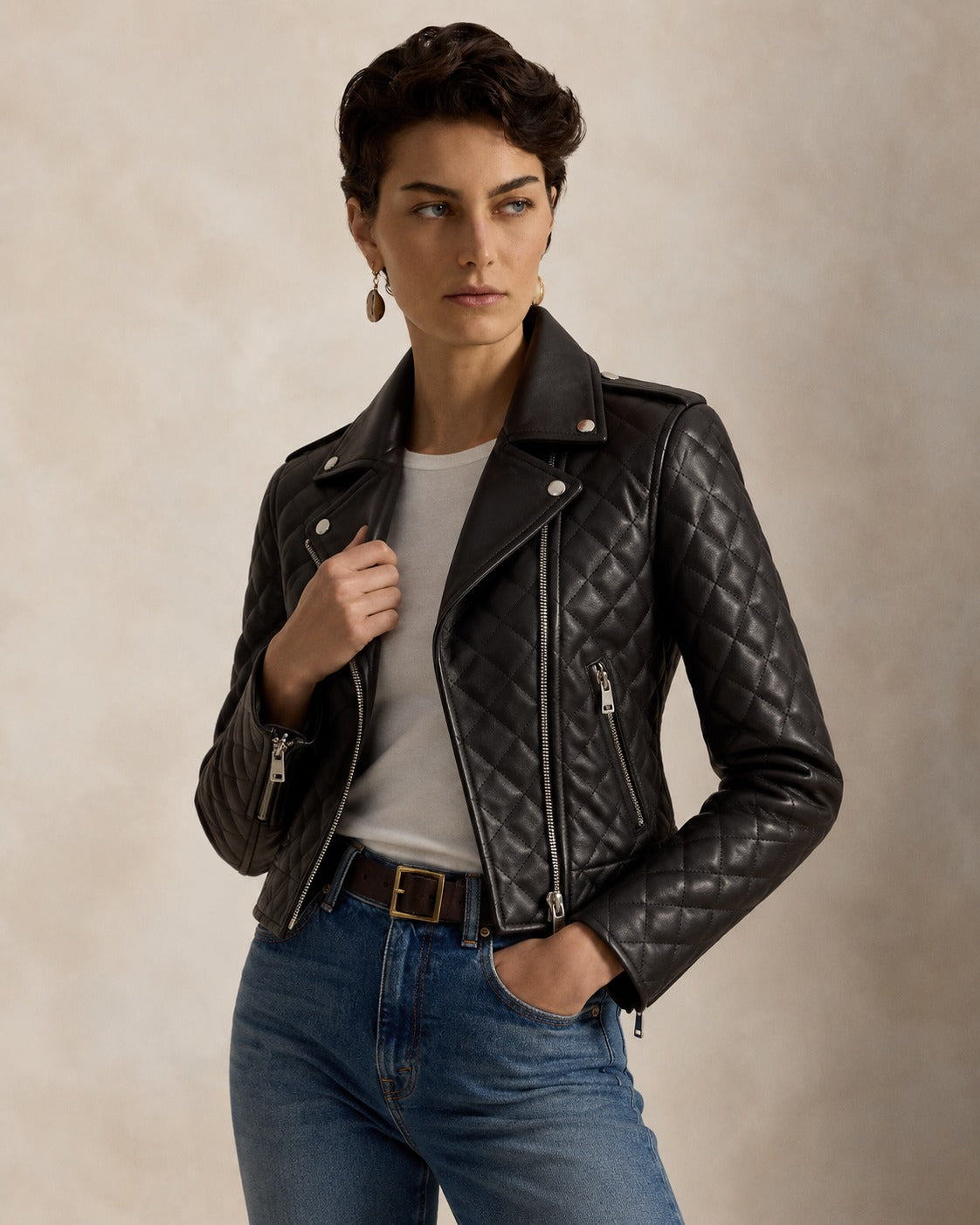 A woman wearing a black quilted leather moto jacket over a white t-shirt and blue jeans, standing against a textured beige studio background.