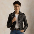 A model wearing a black quilted leather moto jacket over a white t-shirt and blue jeans, standing against a textured beige background.