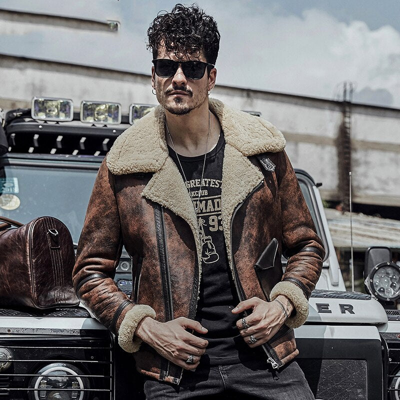 Man wearing a brown leather jacket with fur lining, sunglasses, and a black t-shirt with graphics, standing in front of a vehicle.