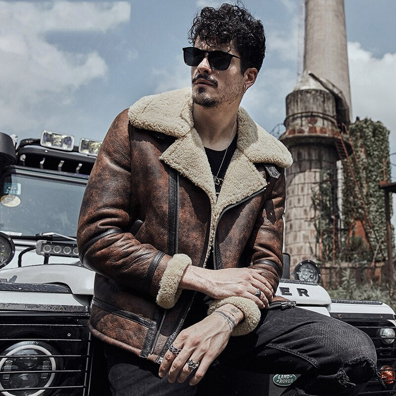 Man wearing a brown shearling jacket and sunglasses sitting in front of a vehicle with a industrial background.