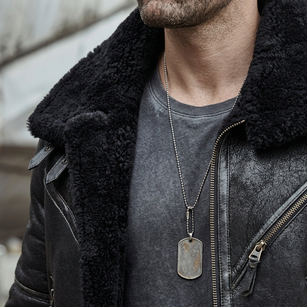 A close-up of a man wearing a black shearling-lined leather jacket over a dark gray t-shirt, featuring a silver-toned dog tag necklace.