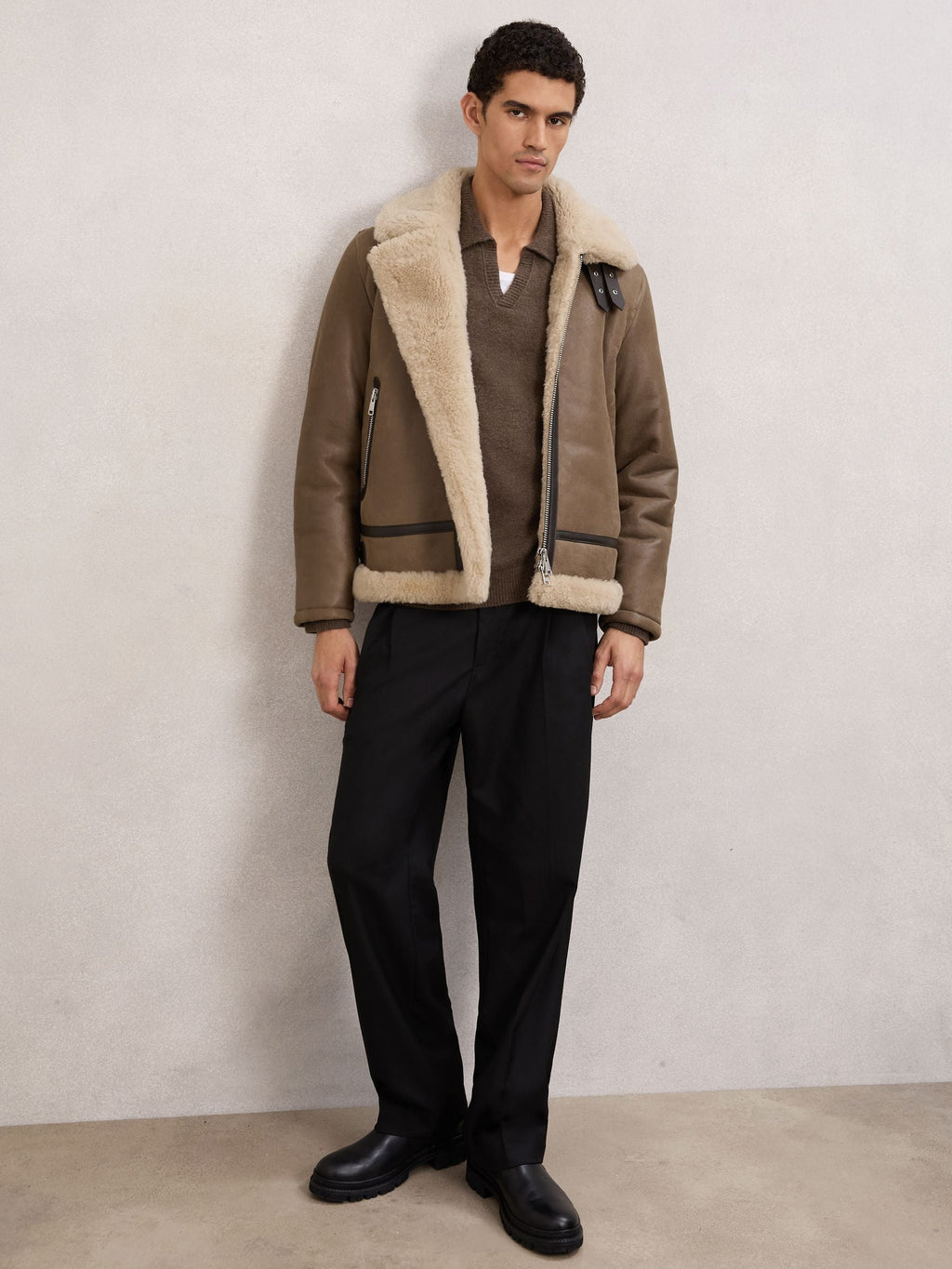 A man wearing a brown shearling-lined aviator jacket over a brown sweater and black trousers, standing against a textured light gray wall.