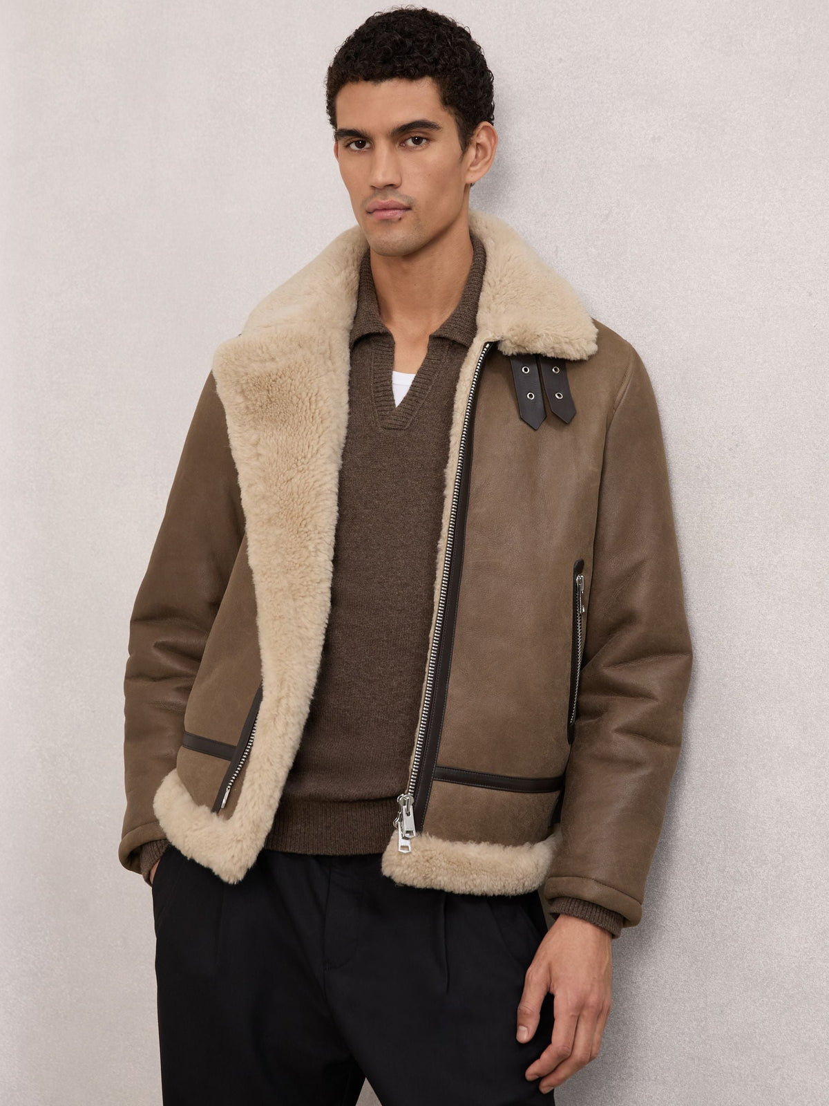 A man wearing a brown leather aviator-style jacket with a thick cream shearling collar and lining, layered over a brown sweater and black trousers, standing against a textured light gray wall.