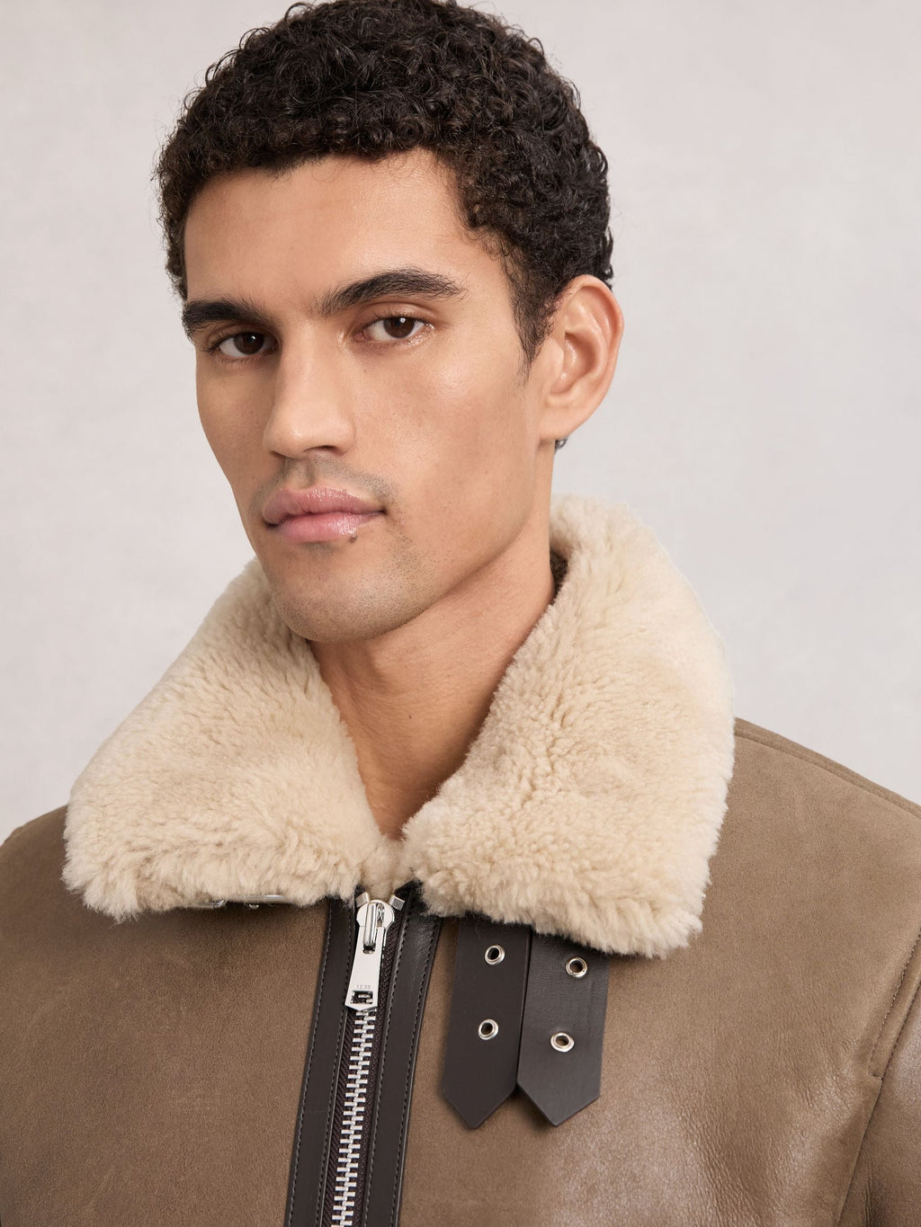 A close-up of a man wearing a brown leather aviator-style jacket with a thick cream shearling collar and a black zipper detail.