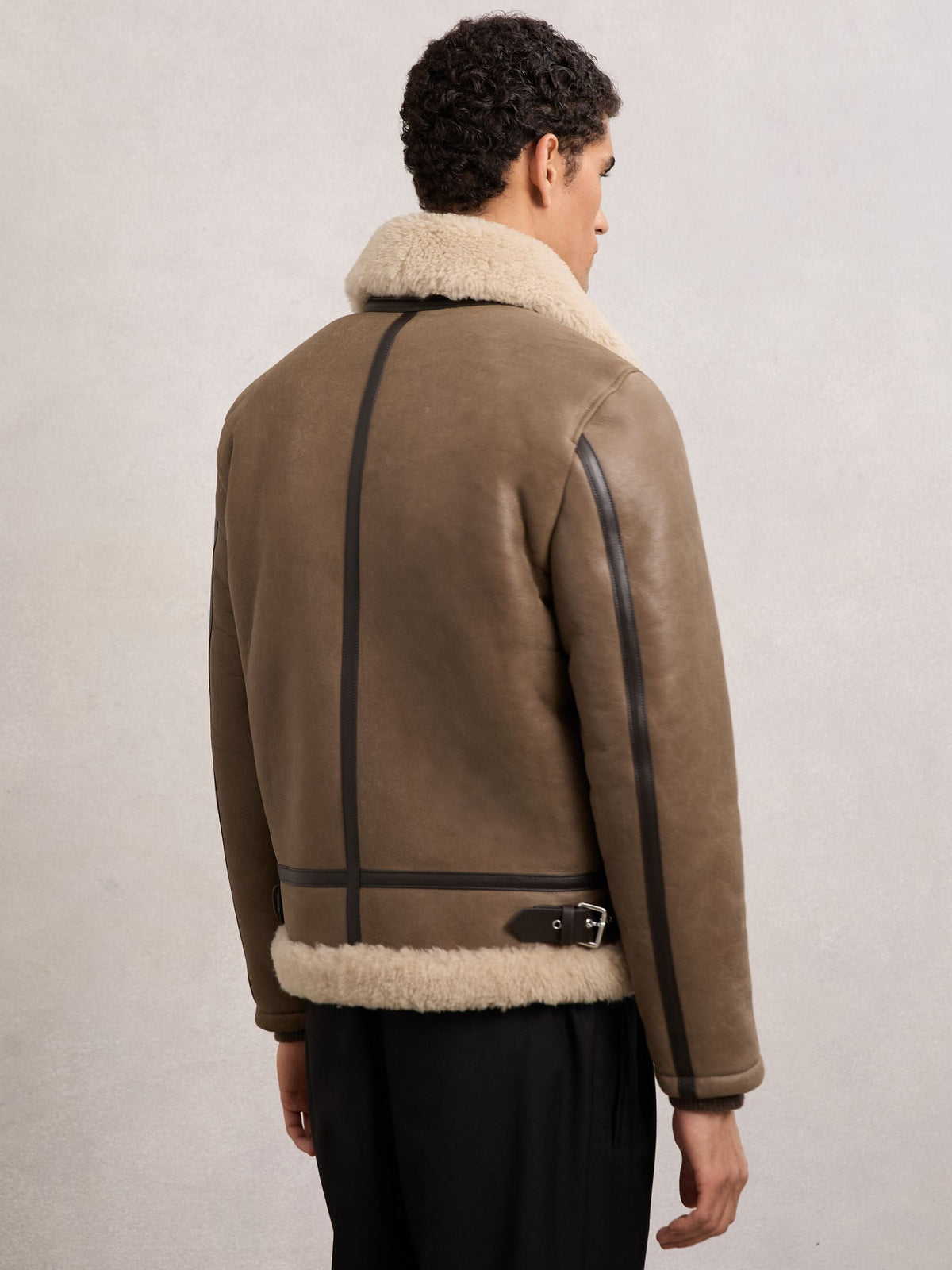 A man shown from the back wearing a brown shearling-lined aviator jacket with black trim and a beige collar, paired with black trousers, against a neutral light gray background.