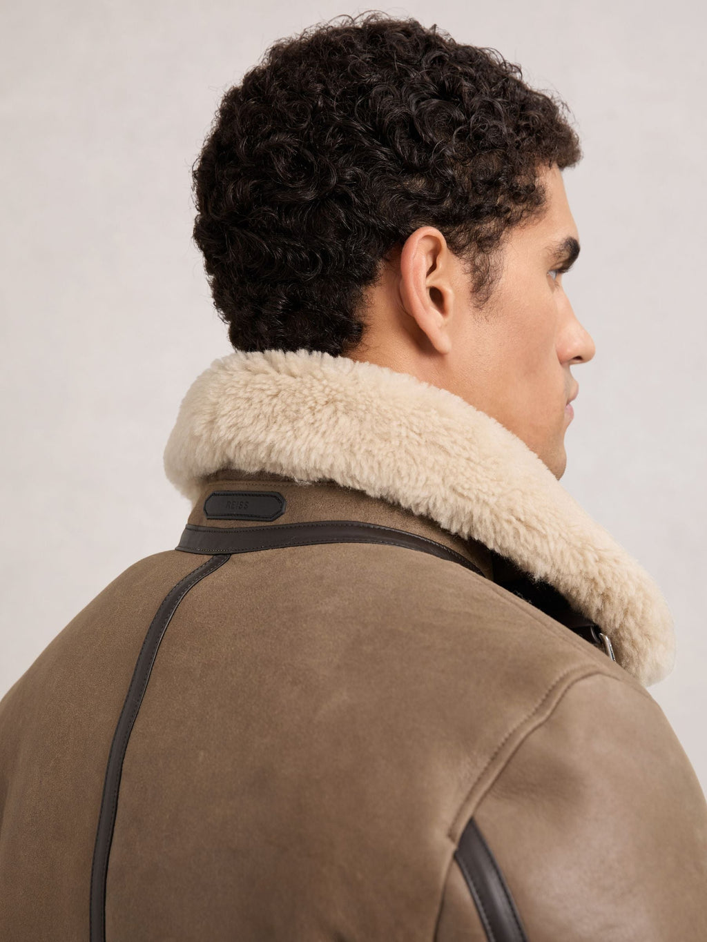 A man shown from the back and side wearing a brown shearling-lined jacket with a thick cream-colored collar and dark leather trim, against a light gray studio background.