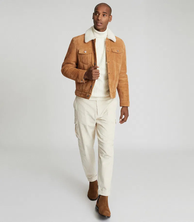 A man modeling a tan suede jacket with a white sherpa collar, worn over a white turtleneck sweater, off-white cargo pants, and brown suede boots, against a light gray studio background.