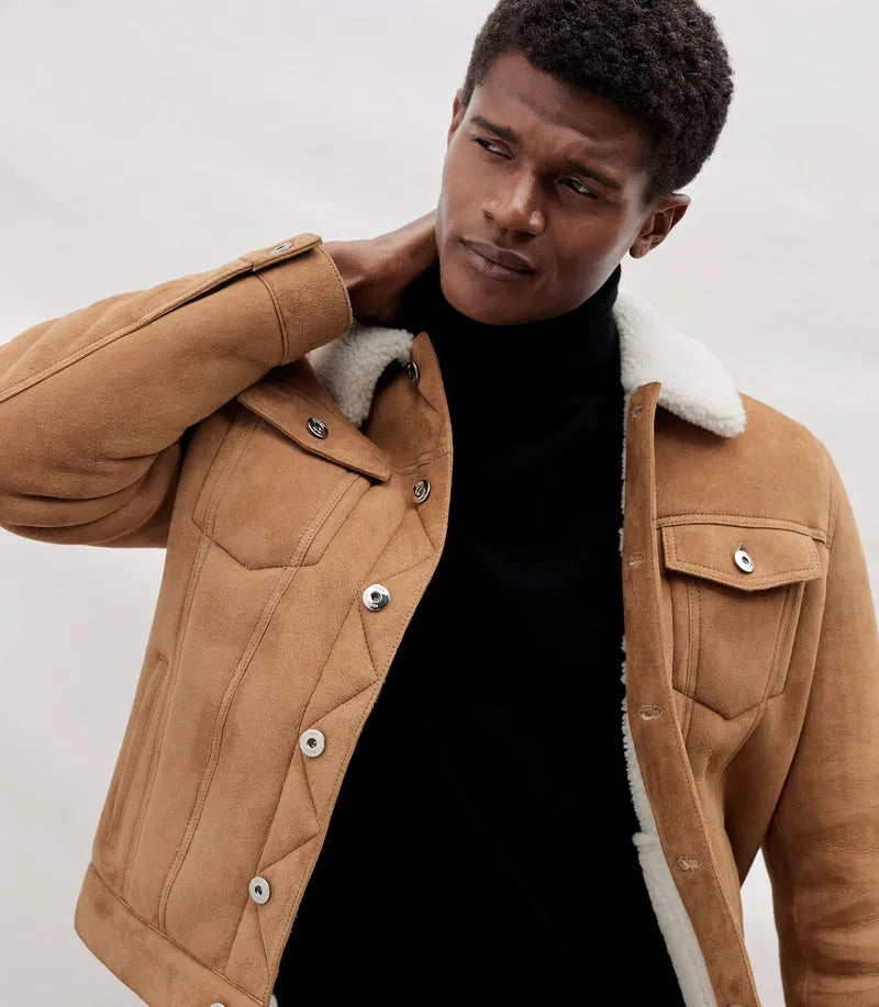 A man wearing a tan suede jacket with a white shearling lining over a black turtleneck sweater, photographed against a light gray studio background.