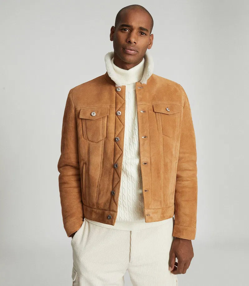 A man wearing a tan suede jacket over a cream cable-knit turtleneck sweater and cream trousers, photographed against a light gray studio background.