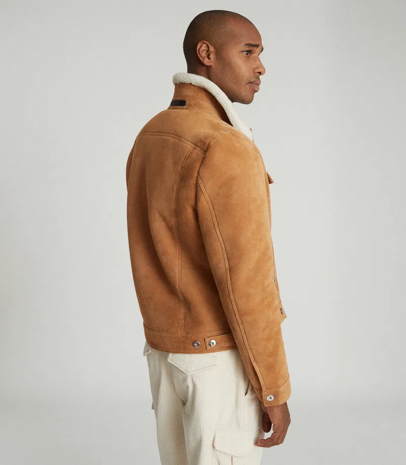 A man shown in profile wearing a tan suede jacket with a white shearling-style collar and off-white pants, photographed against a plain light gray studio background.