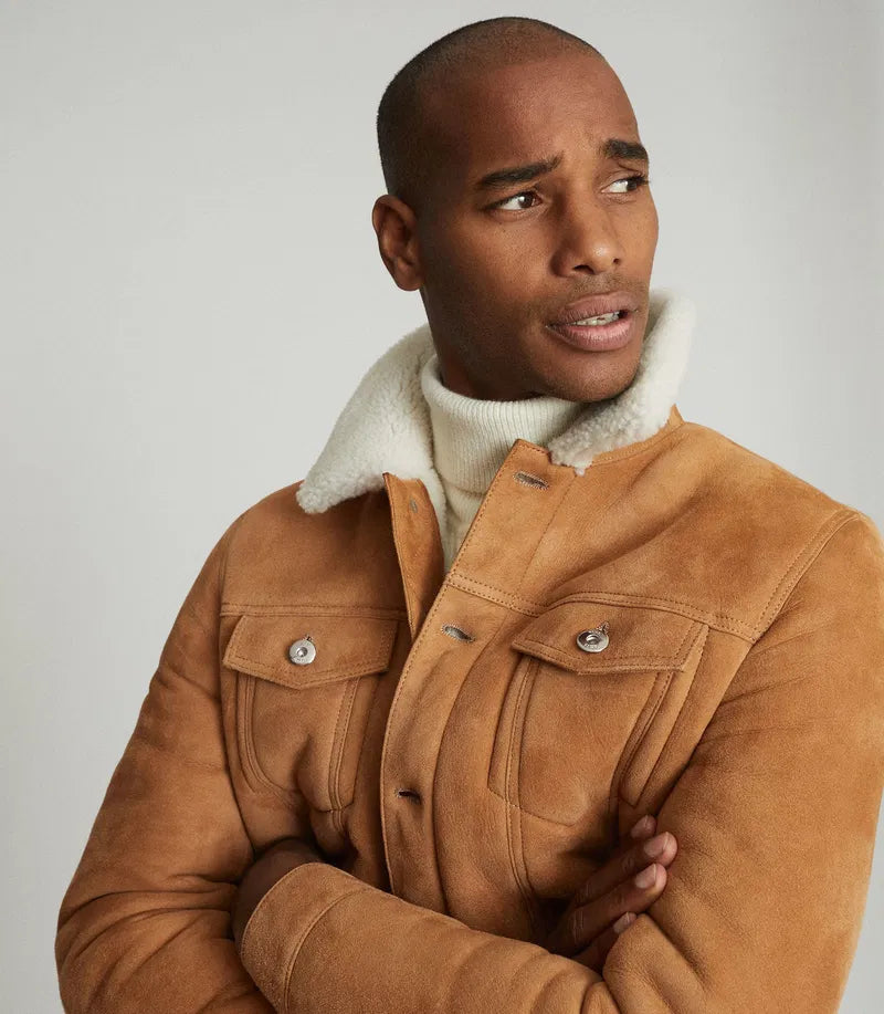 A man wearing a tan suede jacket with a white shearling-style collar over a white turtleneck sweater, photographed against a plain light gray studio background.