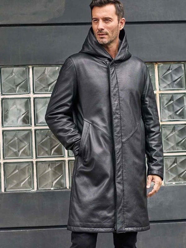 Man wearing a black leather coat standing in front of a glass block wall.