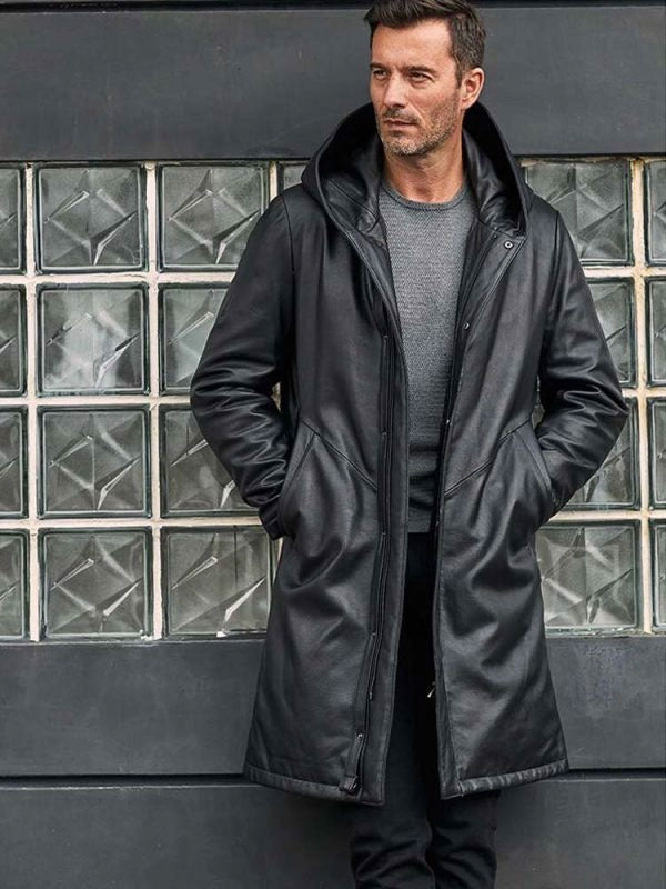 Man wearing a black coat standing against a glass block wall.