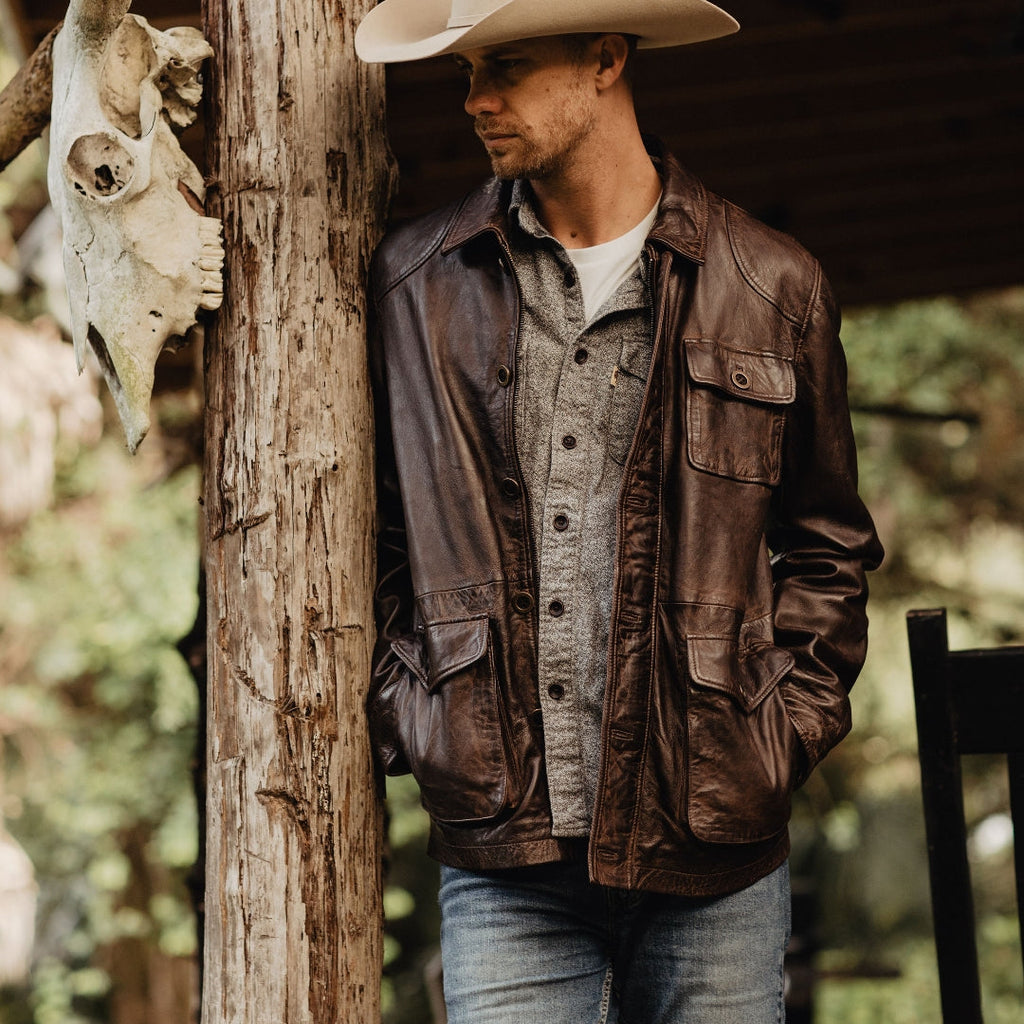 Man wearing a cowboy hat and leather jacket leaning against a wooden post with a blurred natural background