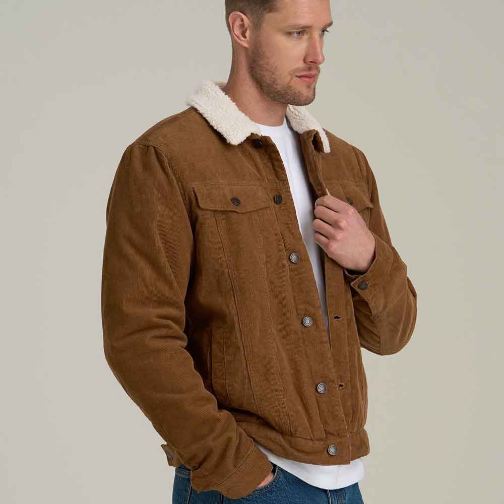 A man wearing a brown corduroy jacket with a white sherpa collar, layered over a white t-shirt and blue jeans, photographed against a solid light gray background.