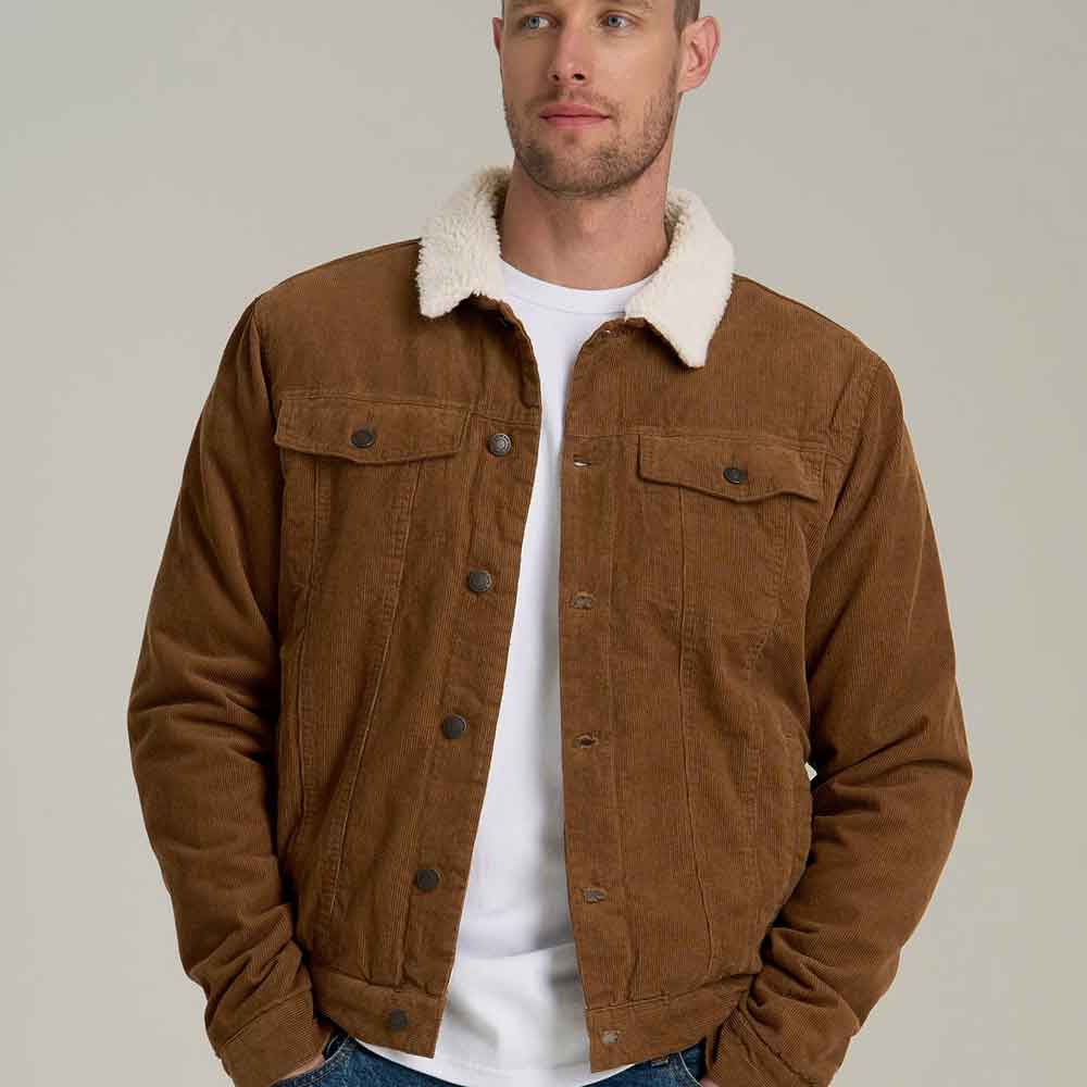 A man wearing a brown corduroy trucker jacket with a white sherpa collar, layered over a white t-shirt and blue jeans, against a plain light gray studio background.