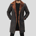 A man wearing a long dark brown leather-like coat with a warm brown faux-fur collar and lining, layered over a black shirt and pants, against a solid gray background.