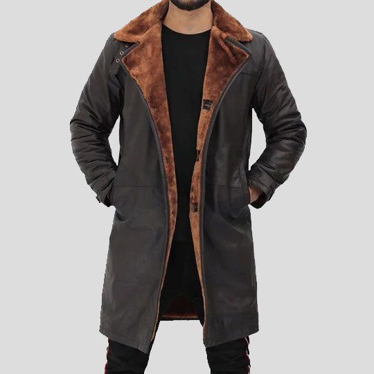 A man wearing a long dark brown leather-like coat with a warm brown faux-fur collar and lining, layered over a black shirt and pants, against a solid gray background.