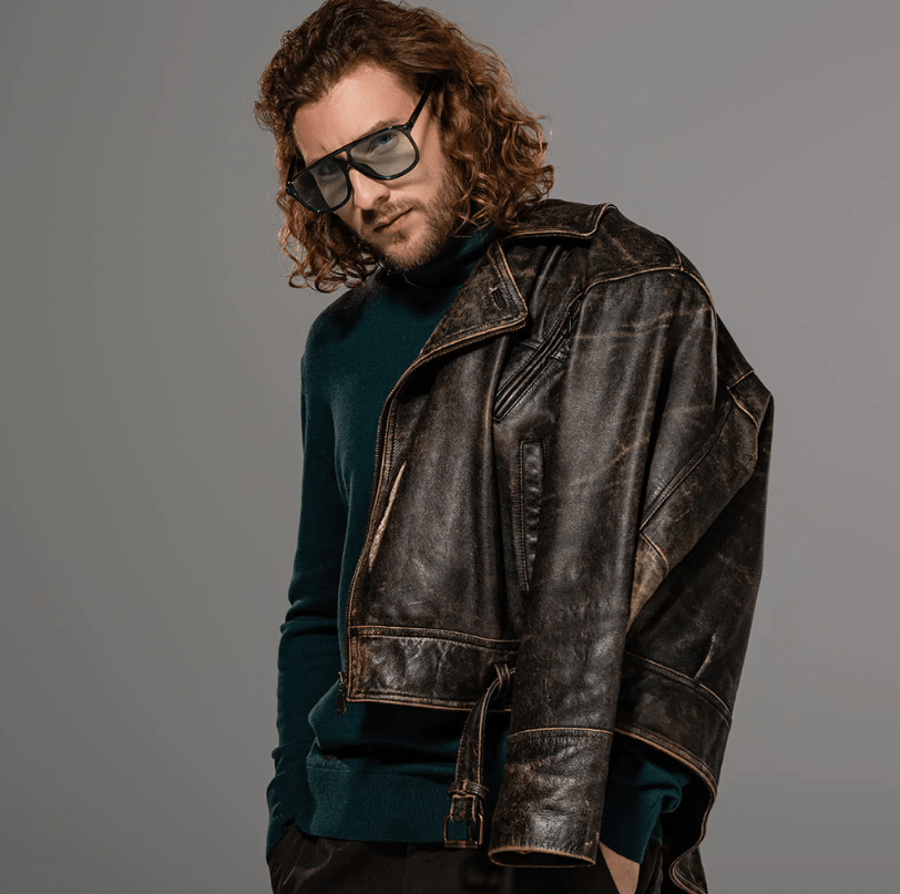 A man with long curly brown hair and sunglasses wearing a distressed dark brown leather jacket over a dark teal sweater, posed against a solid gray studio background.