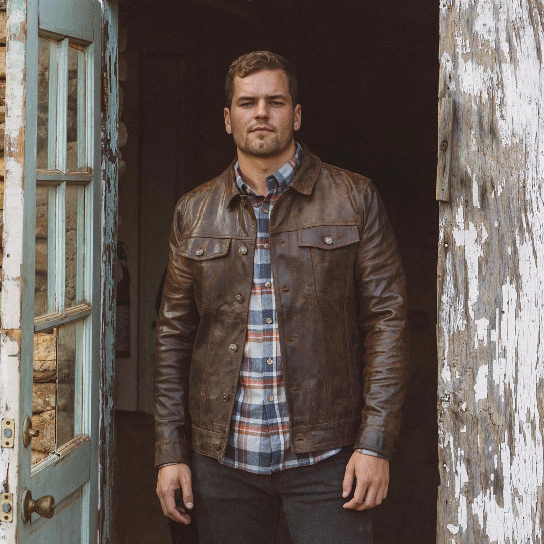 Man wearing a brown leather jacket and plaid shirt standing in a doorway.
