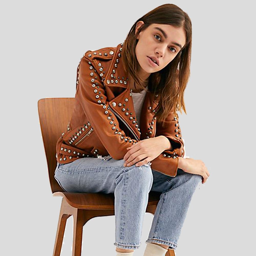 Woman wearing a brown studded leather jacket and blue jeans sitting on a wooden chair.