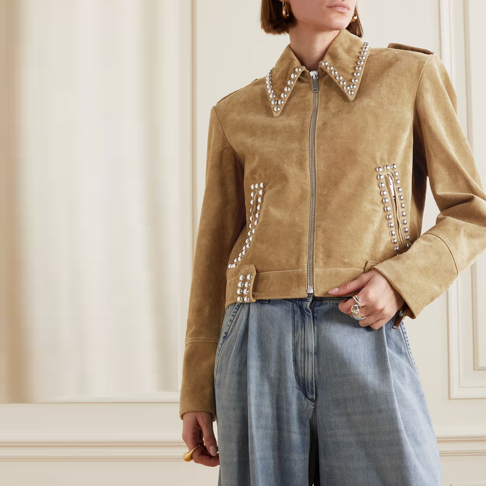 A cropped suede jacket in tan with silver-tone studs and pearl-like embellishments on the collar and front pockets, worn by a model with blue jeans in an elegant indoor setting.
