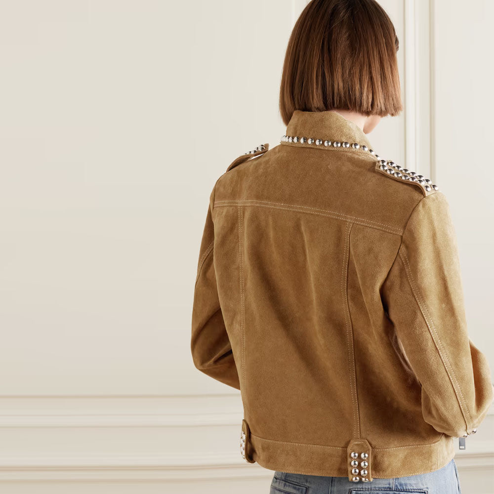 A woman shown from the back wearing a tan suede jacket with silver-tone studs on the shoulders and hem, paired with blue jeans against a cream-colored wall.