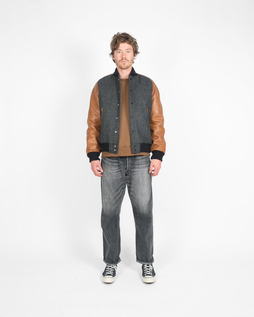 Man wearing a gray jacket with brown sleeves, brown shirt, and gray jeans on a white background