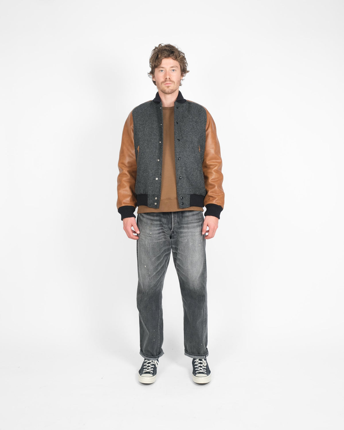 Man wearing a gray jacket with brown sleeves, brown shirt, and gray jeans on a white background